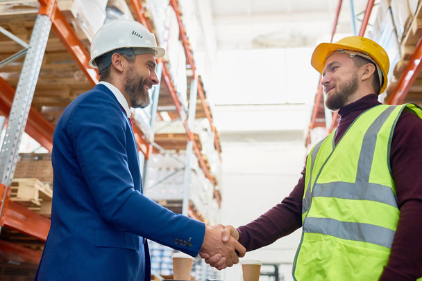 Waist up portrait of handsome mature businessman shaking hands with worker wearing hardhat standing against warehouse shelves in background - Photo, Image