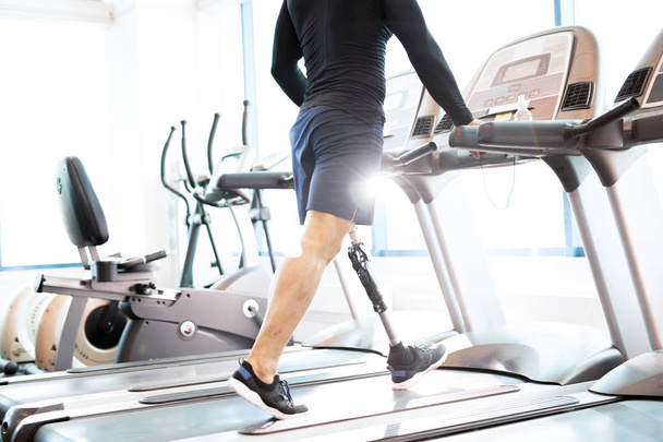 Niedrigwinkel-Porträt eines bis zur Unkenntlichkeit muskulösen Mannes mit Beinprothese beim Laufen auf dem Laufband während des Trainings in einem modernen, sonnenbeschienenen Fitnessstudio - Foto, Bild