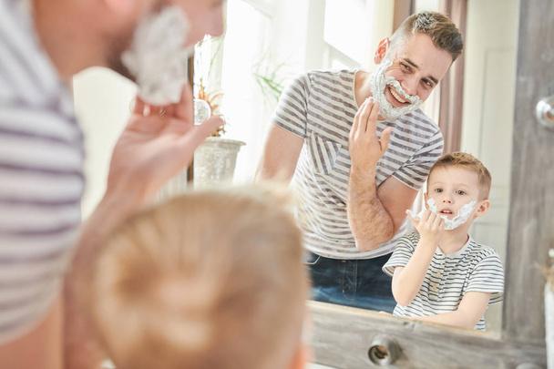 Teplé tónovaný portrétu pohledný tatínek učí roztomilé malého syna jak se holit, stojící proti zrcadlo s tváře pokryté pěnou v moderní koupelně - Fotografie, Obrázek