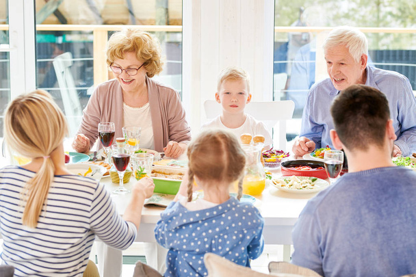 Portrait of happy family enjoying dinner together sitting round festive table with delicious dishes during holiday  celebration, focus on two grandparents and little boy lit by sunlight, copy space - Фото, изображение