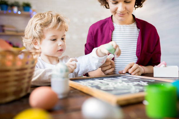 Retrato de lindo niño extendiéndose para escribir en pizarra mientras hace decoraciones de Pascua con la madre
 - Foto, Imagen