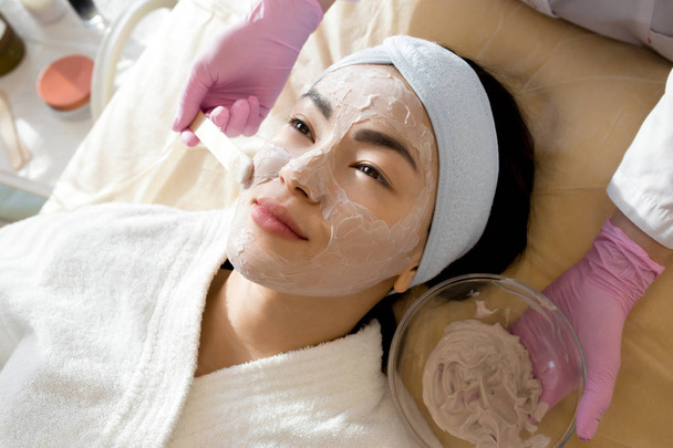 Alto ângulo de perto de cosmetologista irreconhecível aplicando máscara facial no rosto de bela jovem deitada na mesa de massagem durante o tratamento de beleza no salão SPA
 - Foto, Imagem