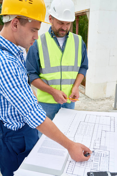 Side view portrait of two construction workers discussing floor plans and engineering documentation on site, pointing at it - Photo, Image