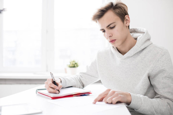 Handsome Caucasian teenage boy in hoodie sitting at desk in library and doing assignment in his notepad - Photo, image