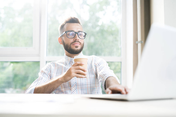 Portret van moderne bebaarde zakenman, werkzaam bij de balie in kantoor met behulp van laptop en genieten van kopje koffie, kopie ruimte - Foto, afbeelding