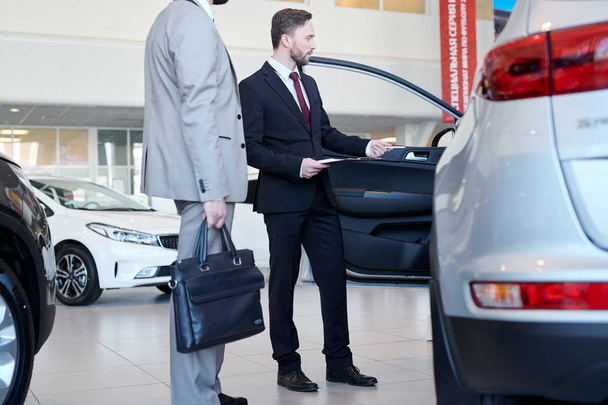 Car salesman representing new car to male customer at auto showroom - Фото, изображение