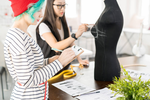 Taille portret van twee hedendaagse creatieve jonge vrouwen werken in modevormgeving en samenwerken inzake project status door kleermakers tabel in moderne atelier studio - Foto, afbeelding
