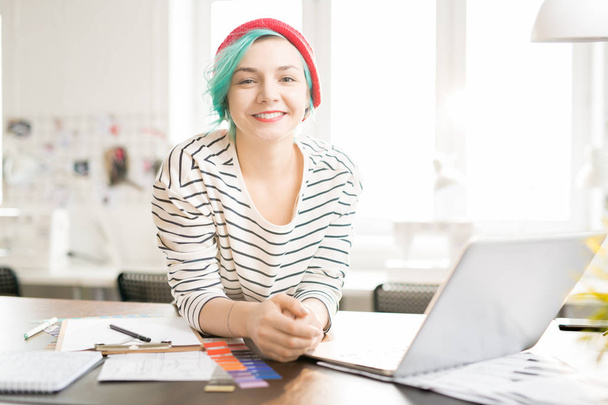 Retrato de una joven creativa contemporánea con el pelo de color menta sonriendo alegremente mirando y la cámara mientras está de pie en la mesa de trabajo en el estudio de diseño iluminado por el sol
 - Foto, Imagen