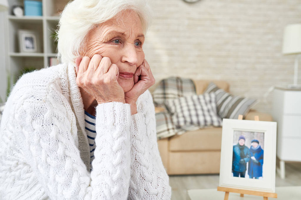 Портрет одинокой пожилой женщины, смотрящей в космос со слезами на глазах, сидящей рядом с фотографией своей семьи, копирующей пространство (фото в рамке от меня
) - Фото, изображение