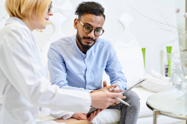 Handsome mixed-race patient answering questions of female physician while she filling in medical record, interior of modern office on background - Foto, afbeelding