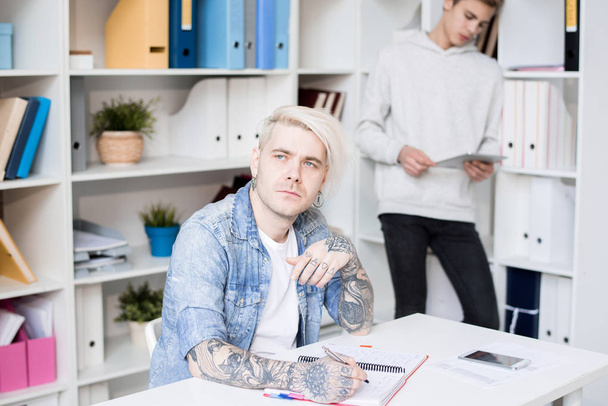 Caucasian adult student with tattoos and dyed hair looking aside pensively while preparing for exam in study room - Fotografie, Obrázek