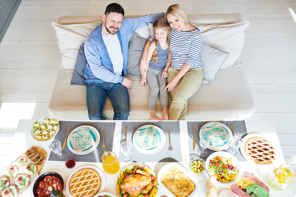 Высокоугольный портрет успешной семьи, наслаждающейся совместным ужином в современной квартире, сидящей на диване с восхитительной маленькой девочкой, все счастливо улыбающейся
 - Фото, изображение