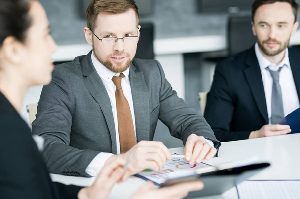 Portrait of successful young businessman talking to colleagues while sitting at meeting table in conference room - Photo, Image