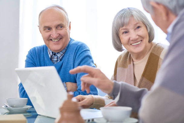 fröhliche ältere Patienten im Betreuten Wohnen teilen sich Laptop, während sie am Tisch chillen und Tee trinken.  - Foto, Bild