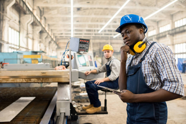 全体的に身に着けているアフリカ系アメリカ人の労働者とヘルメットのデジタル タブレット、背景に近代的な工場の広々 とした製造部門のインテリアの助けを借りて機械の運転 - 写真・画像