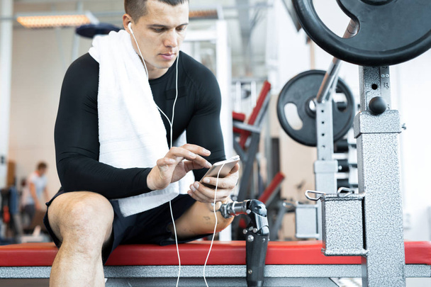 Porträt eines gut aussehenden, muskulösen Mannes mit Beinprothese, der auf einer Bank sitzt und nach dem Training in einem modernen Fitnessstudio entspannt Musik hört, Kopierraum - Foto, Bild