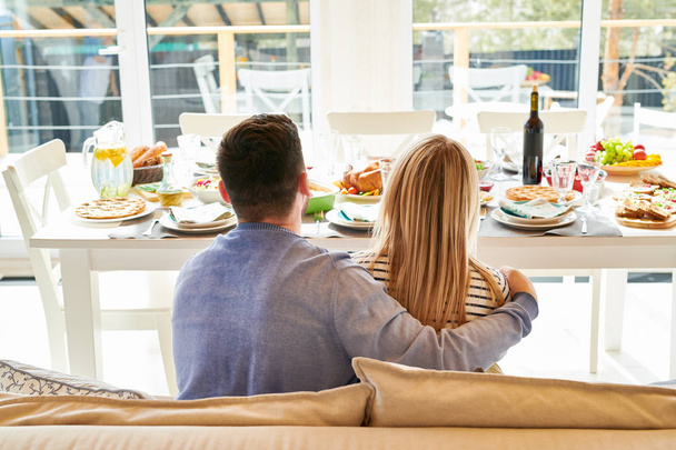 Портрет успешной молодой пары, сидящей на диване за праздничным обеденным столом с видом на окна от пола до потолка дома, копировальное пространство
 - Фото, изображение