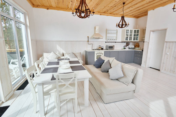 Fotografia de ângulo largo da moderna sala de estar aberta com mesa de madeira branca e balcões de cozinha cinza em design escandinavo minimalista iluminado pela luz solar, espaço de cópia, ninguém
 - Foto, Imagem