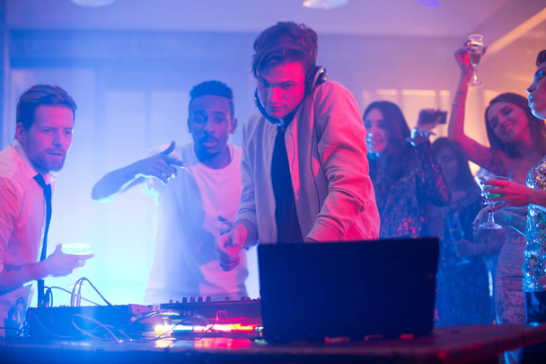 Talentoso joven DJ mezclando música con equipo profesional mientras grupo de amigos alegres bailando y bebiendo champán, interior de club nocturno en el fondo
 - Foto, Imagen