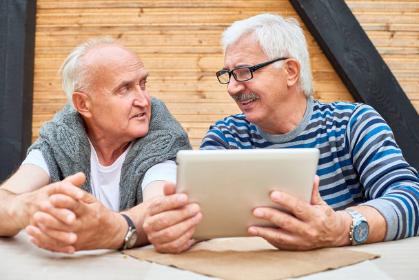 Retrato de dois homens idosos alegres usando tablet digital sentado à mesa no terraço ao ar livre e conversando, espaço de cópia
 - Foto, Imagem