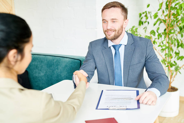 Portrait of handsome business manager shaking hands with female client or partner over contract lying on table after successful negotiations during meeting in modern office, copy space - Photo, Image