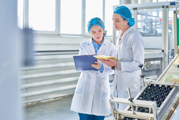 Portrait of  two young female workers wearing lab coats and hair covers standing by  power units and writing on clipboard  in clean production workshop, copy space - Photo, Image