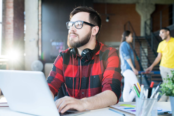 Portrait of modern bearded man wearing glasses and red shirt using laptop looking away pensively in sunlit office of IT developers team, copy space - Photo, Image