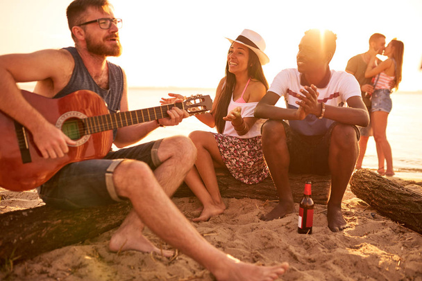 Весело восторженный красивый бородатый молодой человек в очках сидит на бревне и поет песню для многонациональной пары, играя на гитаре с закрытыми глазами на пляже
 - Фото, изображение