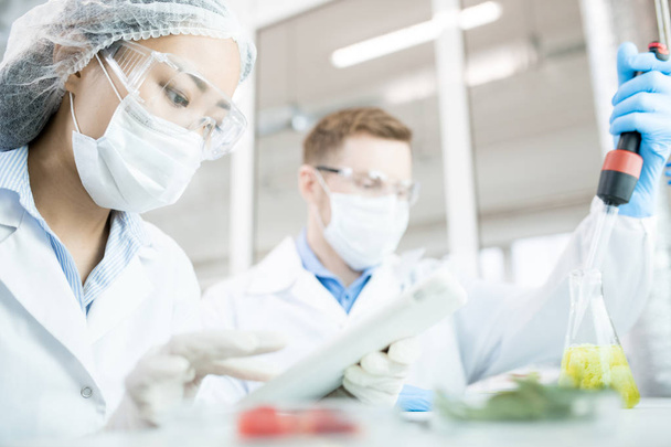 Ritratto di due scienziati moderni che indossano maschere che lavorano alla ricerca alimentare studiando liquidi in becher mentre siedono a tavola in laboratorio
 - Foto, immagini