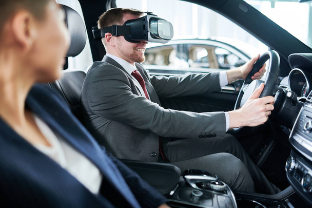 Vista de perfil de cliente potencial alegre sentado en el asiento del conductor y sometido a prueba de conducción de coche nuevo con ayuda de auriculares VR, distribuidor bastante mirándolo
 - Foto, imagen