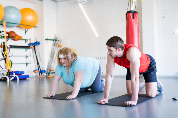 Ganztägiges Porträt einer übergewichtigen Frau bei Fitnessübungen im Fitnessstudio mit Personal Trainer, der ihr hilft - Foto, Bild