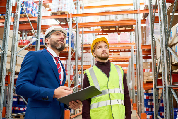 Waist up portrait of warehouse manager holding clipboard talking to worker wearing hardhat and reflective jacket while discussing stock inventory - Photo, Image