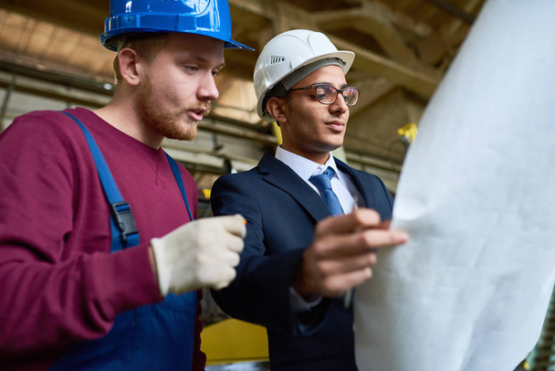 Retrato de bajo ángulo del trabajador de la fábrica hablando con el ingeniero de Oriente Medio sosteniendo planos
 - Foto, imagen