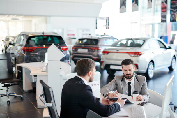 Bearded young male customer signing a contract for rent a car while sitting at car showroom - Photo, Image