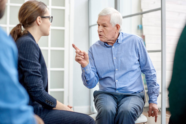 Portret shot van senior patiënt communiceren met zeer professionele psycholoog terwijl je deelneemt groepstherapie sessie, interieur van psychotherapie office op achtergrond - Foto, afbeelding