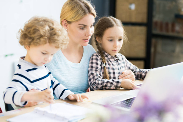 Красивая мать сидит за столом вместе со своей маленькой дочерью и сыном и смотрит на ноутбук
 - Фото, изображение