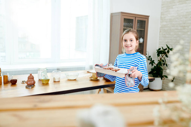 Portraitaufnahme des süßen kleinen Mädchens, das ein Blech mit frisch zubereiteten Cupcakes in der Hand hält und in die Kamera schaut, während sie eine Überraschung für den Muttertag macht - Foto, Bild