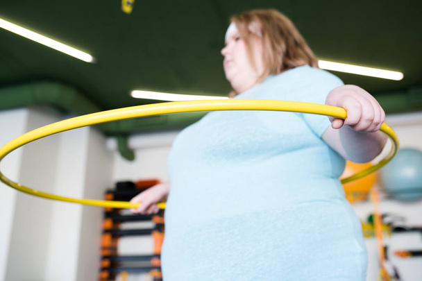 Портрет молодой ожирённой женщины, тренирующейся с хула-хупом в фитнес-клубе, акцент на переднем плане
 - Фото, изображение