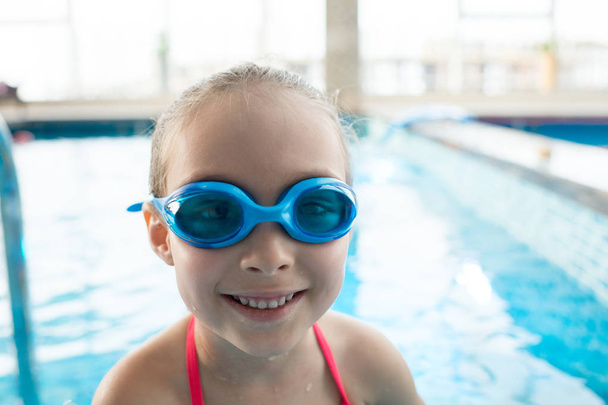 Chica bonita alegre activa en gafas de natación sonriendo a la cámara en la moderna piscina cubierta con agua limpia
 - Foto, imagen
