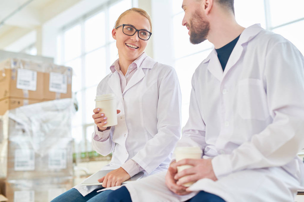 Twee technologen dragen witte jassen zit op ruime verpakkingsafdeling van melkfabriek en genieten van geurige koffie - Foto, afbeelding