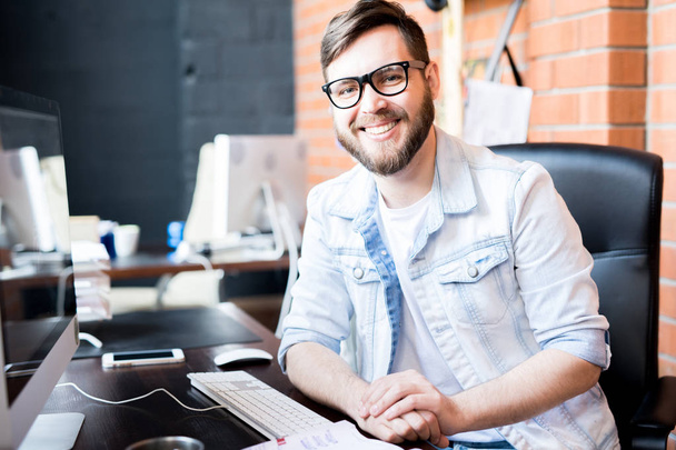 Portrait of contemporary business manager wearing casual clothes smiling happily at camera while sitting at computer desk in modern office - 写真・画像
