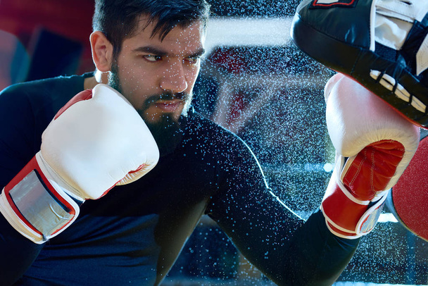 Bearded determined man boxing on ring with trainer while punching focus mitts in water drops.  - Photo, Image