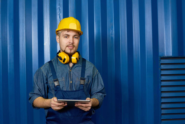 Retrato de la cintura hacia arriba del hombre joven usando hardhat posando contra la pared azul en la fábrica y mirando a la cámara, sosteniendo la tableta digital, espacio de copia
 - Foto, imagen