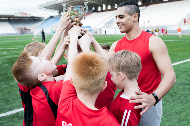 Portrait of junior football team holding trophy together and cheering after winning match in outdoor stadium - Photo, Image