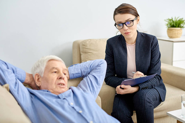 Przygnębiony starszego mężczyzny leżącego na wygodną sofę urzędu psychoterapii i rozmawia z wysoce profesjonalny psycholog młody, ona niezbędnych notatek - Zdjęcie, obraz