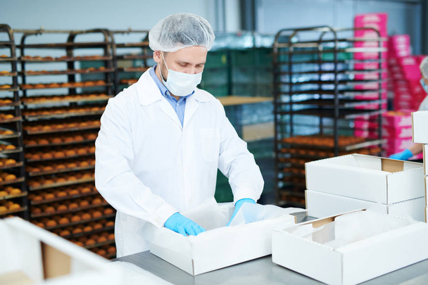 Ouvrier d'usine de confiserie en manteau blanc mettant du parchemin de cuisson dans une boîte d'emballage vide
. - Photo, image