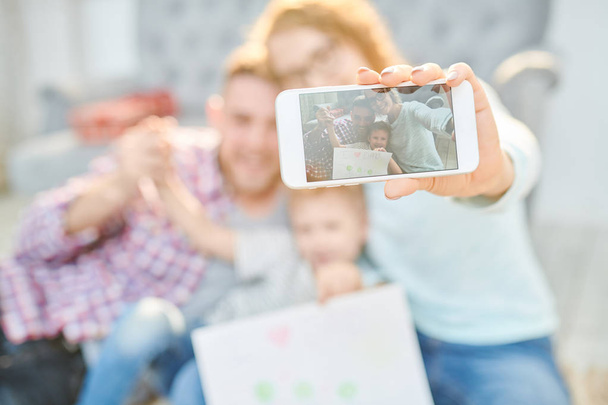 Warm toned portrait of loving young family with cute little son playfully taking selfie at home   in sunlight, focus on picture in smartphone screen, copy space - Foto, Imagen