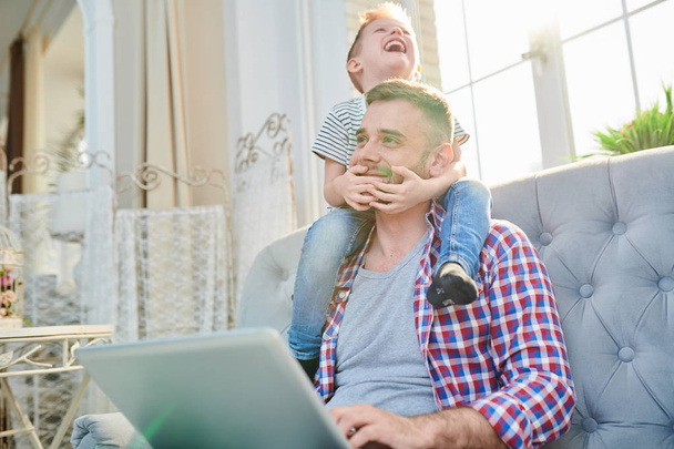 Onvergetelijke momenten van de kindertijd: lachen jongetje zittend op de schouders van zijn liefdevolle bebaarde vader terwijl hij surfen op Internet op laptop, woonkamer interieur op achtergrond - Foto, afbeelding