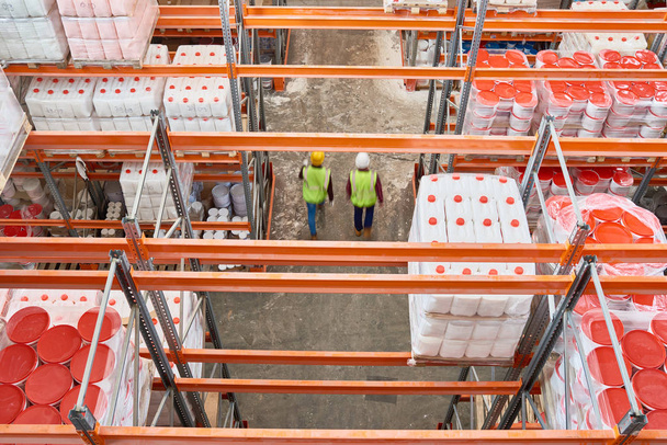 Visão superior imagem de fundo de prateleiras altas em armazém moderno com dois trabalhadores usando chapéus duros andando no corredor, espaço de cópia
 - Foto, Imagem
