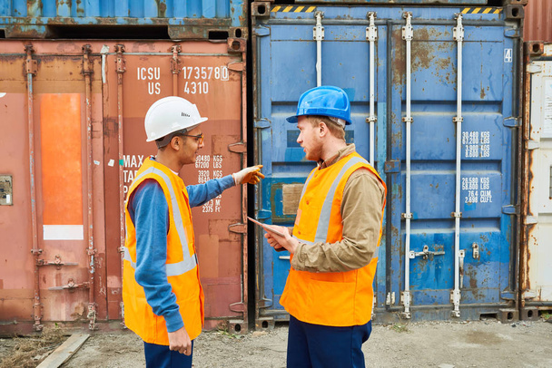 Side view portrait of two dock workers wearing hardhats talking to each other standing against cargo containers - Photo, Image